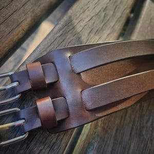 May include: A brown leather watch cuff with two silver buckles. The cuff has two parallel straps and two smaller straps for the buckles. The leather has a textured appearance.