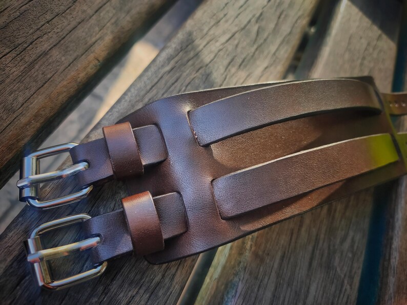May include: A brown leather watch cuff with two silver buckles. The cuff has two parallel straps and two smaller straps for the buckles. The leather has a textured appearance.