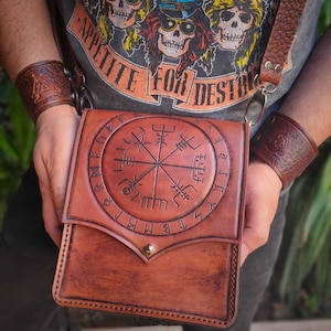 Może przedstawiać: Brązowa skórzana torba na ramię z wytłoczonym okrągłym wzorem. Torba ma zapięcie na klapę z mosiężnym guzikiem. Pasek i pasujące mankiety na nadgarstki są również ze skóry w kolorze brązowym. Osoba ma na sobie szarą koszulkę z napisem "APPETITE FOR DESTRUCTION".