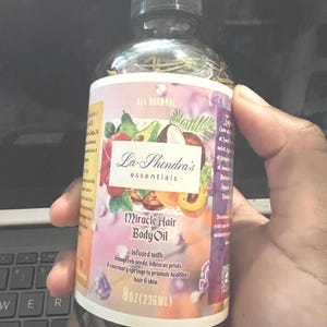May include: A brown glass bottle with a dropper top, labeled "La-Shondra's essentials" and "Miracle Hair Body Oil". The label is pink and purple with a floral design and features a list of ingredients including fenugreek seeds, hibiscus petals, and rosemary springs. The bottle contains 8 oz (236 ml) of oil.