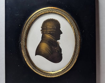 Puede incluir: Retrato de silueta antigua en un marco ovalado. El retrato presenta un perfil de una persona en tonos oscuros, sobre un fondo blanco. El marco ovalado está rodeado por un borde dorado y está dentro de un marco rectangular negro, coronado con un acento dorado decorativo.