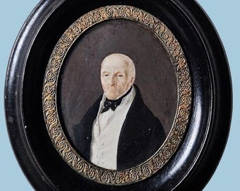 Puede incluir: Un retrato ovalado de una persona calva con expresión seria, que viste camisa blanca, pajarita negra y abrigo oscuro. La obra está enmarcada en madera negra con un borde interior dorado y texturizado con detalles ornamentales.