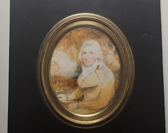 May include: An antique portrait miniature of a man in a yellow coat and white cravat, set within an oval frame. The portrait is mounted on a black rectangular backing with a decorative brass hanger.