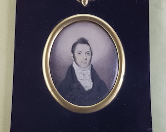 Puede incluir: Un retrato en miniatura de un hombre con un abrigo negro y una corbata blanca, enmarcado en negro con un paspartú ovalado dorado y un remate de bellota dorado.