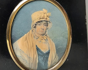 Puede incluir: Un retrato en miniatura enmarcado de una mujer que lleva un sombrero amarillo con un lazo y una chal blanca. El retrato está pintado en acuarela sobre marfil y está enmarcado en un marco negro con un paspartú ovalado dorado.
