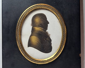Op de afbeelding: Een zwart ingelijst silhouetportret van een persoon in profiel, met een gouden ovale lijst en een eikelvormige ophanglus.