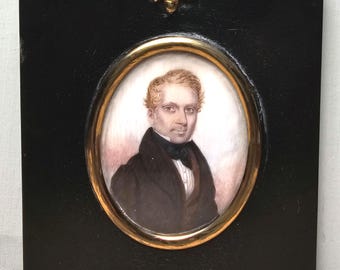Puede incluir: Un retrato en miniatura de un hombre con un abrigo negro y una camisa blanca, pintado sobre marfil y enmarcado en un marco de madera negro con un marco ovalado dorado y un remate de bellota.