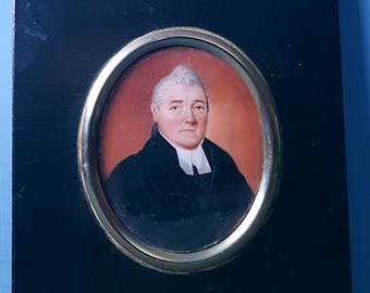 Op de afbeelding: Een ingelijst miniatuurportret van een persoon in een zwarte jas en witte kraag. Het ovale portret staat tegen een rode achtergrond, is verguld en gemonteerd op een zwart rechthoekig frame. Een decoratieve metalen eikel en blad sieren de bovenkant.