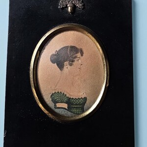 May include: A rectangular black wall frame featuring an oval gold-toned border surrounding a profile portrait of a person with dark hair in a green patterned dress. A silver-toned metal ornament shaped like an acorn and oak leaves sits at the top.
