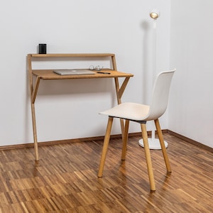 Könnte beinhalten: Ein weißer Stuhl mit Holzbeinen steht neben einem Holzschreibtisch mit einem Laptop, einer Brille und einem schwarzen Behälter darauf. Eine weiße Stehlampe mit weißem Schirm steht im Hintergrund.