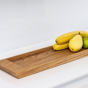 Könnte beinhalten: Ein langes, rechteckiges Holztablett mit erhöhtem Rand. Das Tablett ist aus hellbraunem Holz gefertigt und hat eine natürliche Oberfläche. Das Tablett ist mit gelben Bananen und einer grünen Birne gefüllt.