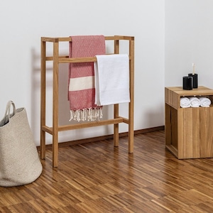 Pode incluir: Um suporte de toalha de madeira com duas barras para pendurar toalhas. O suporte é feito de madeira natural e tem um design simples e moderno. Uma toalha branca e uma toalha listrada vermelha e branca estão penduradas no suporte. Um pequeno banquinho de madeira com uma prateleira está no fundo.