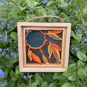 Può includere: Un acchiappasogni incorniciato con piume marroni, arancioni e gialle. L'acchiappasogni è blu e marrone su uno sfondo bianco. La cornice è in legno marrone chiaro.