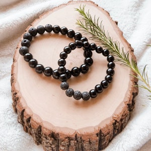 May include: Two black beaded bracelets, one with smooth beads and the other with textured lava rock beads. The bracelets are displayed on a rustic wooden slice with a sprig of greenery.