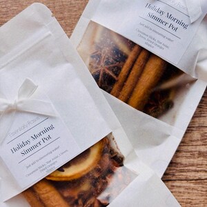 May include: Two white pouches with a clear window showing the contents inside. The pouches are labeled "Holiday Morning Simmer Pot" and contain cinnamon sticks, star anise, cloves, dried oranges, and allspice. Just add to simmering water!