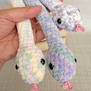 May include: Three plush crochet snakes made from soft, variegated yarn in pastel shades of pink, yellow, blue, purple, and white. Each snake features black plastic bead eyes and a small, pink crocheted tongue. The text smiley tangerine is visible.