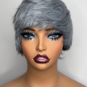 May include: A short, silver-grey wig with a side part and bangs. The wig is styled in a pixie cut with layers and texture.