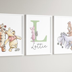 May include: Three white-framed wall art prints featuring Winnie the Pooh characters. The left print shows Tigger and Pooh. The center print displays a large green letter L, Piglet, and the name Lottie. The right print shows Eeyore, Kanga, and Roo.