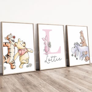 May include: Three framed wall art prints featuring Winnie the Pooh characters. The first shows Tigger and Pooh, the second a large pink letter L with Piglet and the name Lottie, and the third Eeyore, Roo, and Roo's mother with flowers.