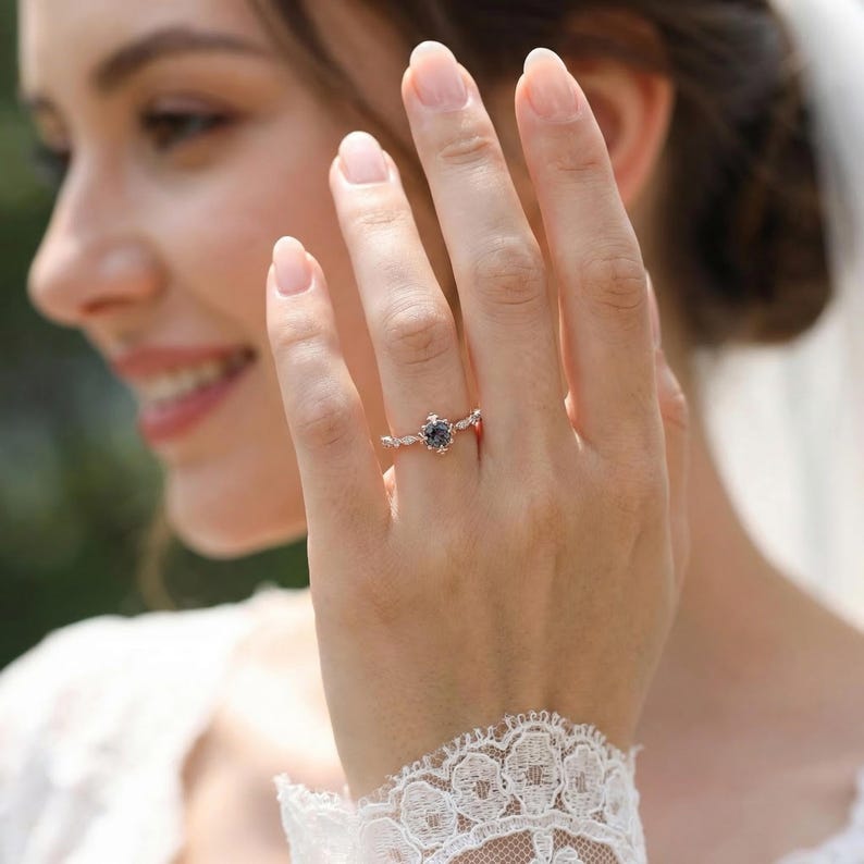 以下が含まれることがあります： 中央に濃い青色の宝石が付いたローズゴールドのリング。リングは、小さなアクセントストーンを備えた繊細なフローラルデザインが特徴です。リングは人の指にあり、白いレースのスリーブが前景にあります。