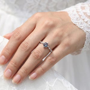 以下が含まれることがあります： 中央に丸い濃い青色の宝石が付いたシルバーリング。リングは、小さな透明なアクセントストーンを備えた、繊細な花にインスパイアされたデザインが特徴です。リングは指にあり、背景には白いレースのスリーブがあります。