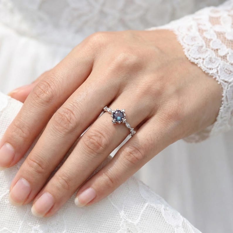以下が含まれることがあります： 中央に丸い濃い青色の宝石が付いたシルバーリング。リングは、小さな透明なアクセントストーンを備えた、繊細な花にインスパイアされたデザインが特徴です。リングは指にあり、背景には白いレースのスリーブがあります。