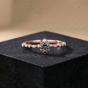 以下が含まれることがあります： ローズゴールドのリング。中央に楕円形のティール色の宝石がセットされています。バンドには小さなクリアなアクセントストーンと装飾的な要素が施されています。リングは暗い表面に置かれ、その複雑なデザインと色を際立たせています。