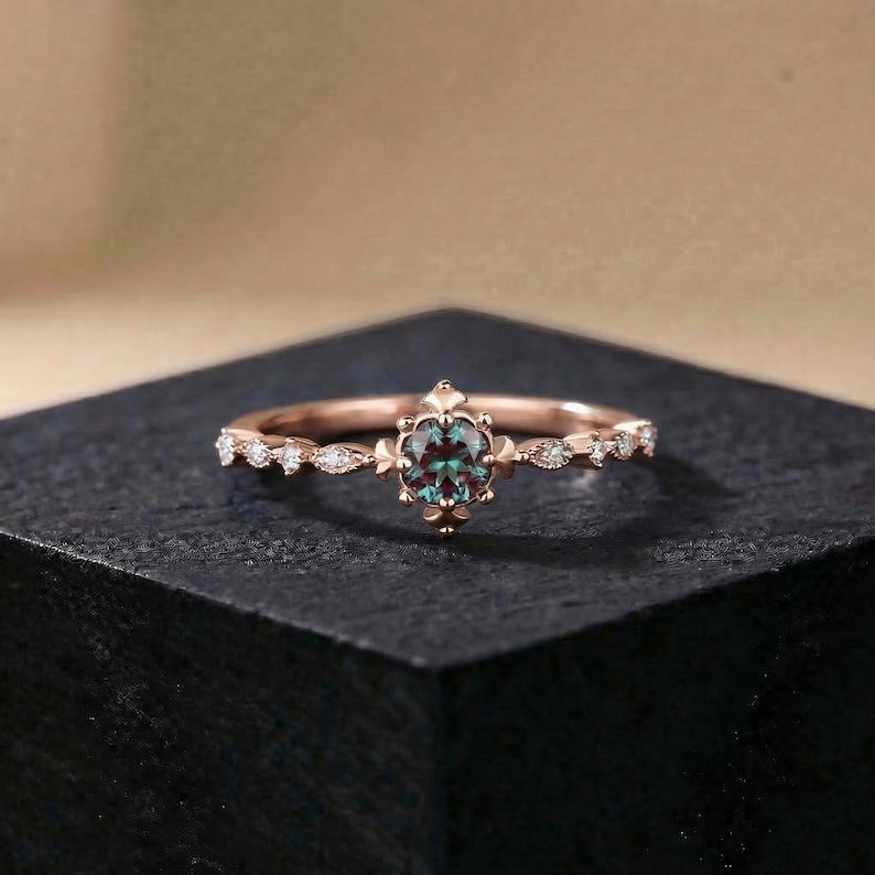 以下が含まれることがあります： ローズゴールドのリング。中央に楕円形のティール色の宝石がセットされています。バンドには小さなクリアなアクセントストーンと装飾的な要素が施されています。リングは暗い表面に置かれ、その複雑なデザインと色を際立たせています。