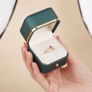 以下が含まれることがあります： 紫色の宝石が付いた金の指輪が、開いたエメラルドグリーンのリングボックスの中に展示されています。ボックスは金のトリムと白い内装です。リングボックスは人の手で持たれています。