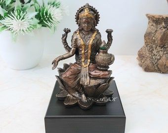 May include: A gold and bronze statue of the Hindu goddess Lakshmi sitting on a lotus flower. She is holding a pot of gold in her right hand and a lotus flower in her left hand. The statue is on a black wooden base.
