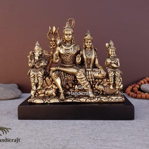 May include: A detailed brass sculpture depicting a Hindu family, including Shiva, Parvati, Ganesha, and Kartikeya. The figures are seated on a textured base, mounted on a black rectangular stand. The sculpture is a religious item.