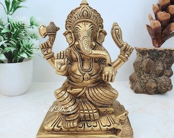 May include: A gold-colored brass statue of Ganesha, the Hindu god of wisdom and prosperity, sitting on a lotus base. The statue is intricately detailed and features Ganesha with his elephant head, four arms, and a broken tusk.