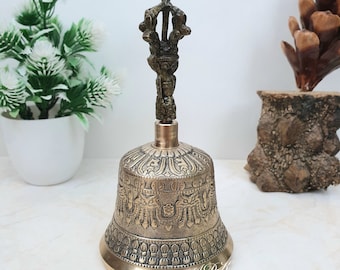 Può includere: Una campana dorata con intagli intricati e un manico decorativo. La campana è appoggiata su una superficie bianca.
