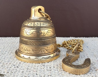 Può includere: Una campana in ottone dorata con intricati disegni incisi è appesa a una catena con un gancio. La campana ha una forma arrotondata e una piccola apertura nella parte superiore. La catena è fatta di anelli metallici collegati.