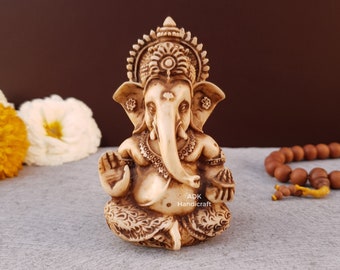 Puede incluir: Una pequeña estatua de color marfil de la deidad hindú Ganesha. La estatua está sentada con las piernas cruzadas y las manos en un gesto de bendición. Ganesha lleva una corona y un collar, y tiene una trompa y orejas grandes. La estatua está sobre un fondo gris con un mala (cuentas de oración) de madera en el fondo.