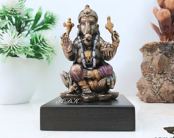 Puede incluir: Una estatua en tonos bronce de la deidad hindú Ganesha, sentado sobre una base de flor de loto, encima de un bloque de madera oscura. La estatua presenta detalles intrincados, incluyendo una corona, joyas y un cuenco de fruta. La base está inscrita con "A.D.K."