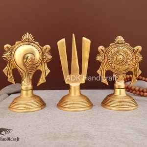 May include: Three gold-colored metal decorative figurines. The figurine on the left is a conch shell, the middle figurine is a trident, and the figurine on the right is a wheel with a flower design. The figurines are on a gray textured surface.