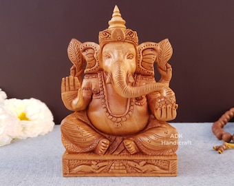 Könnte beinhalten: Eine handgeschnitzte Holzstatue der hinduistischen Gottheit Ganesha. Die Statue ist in einer hellbraunen Farbe bemalt und weist komplizierte Details auf. Ganesha ist sitzend dargestellt, mit erhobenem Rüssel und einem Süßigkeit in der Hand.