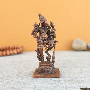 May include: A bronze-colored statue of a deity with a pig-like face, holding a wheel and standing with a smaller figure. The statue is on a square base. The background is a light orange color.