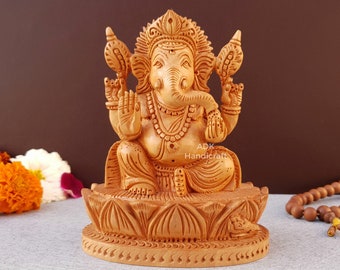 Op de afbeelding: Een handgesneden houten beeld van de hindoe-god Ganesha, zittend op een lotusbasis. Het beeld is geschilderd in een lichtbruine kleur en heeft ingewikkelde details. Het beeld is ongeveer 15 cm hoog.