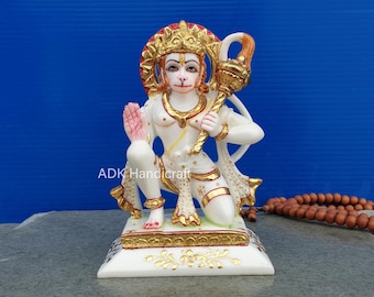 Może przedstawiać: Biała marmurowa statua hinduskiego bóstwa Hanuman, siedząca na białej podstawie z złotymi akcentami. Statua trzyma maczugę w prawej ręce i ma czerwoną i złotą aureolę wokół głowy.