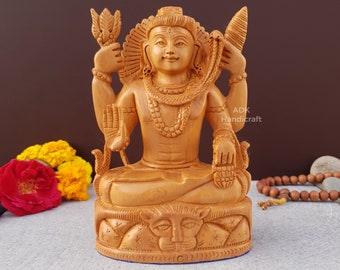 May include: A wooden statue of the Hindu deity Shiva, sitting in a meditative pose with his hands in a mudra. He is adorned with a snake around his neck and a third eye on his forehead. The statue is carved with intricate details and is sitting on a base with a lion's head.