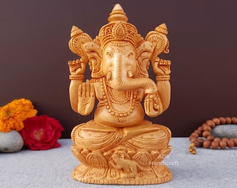 Op de afbeelding: Een handgesneden houten beeld van de hindoe-god Ganesha. Het beeld is geschilderd in een lichtbruine kleur en heeft ingewikkelde details. Ganesha wordt afgebeeld met vier armen, een groot hoofd en een olifantenstam. Het beeld zit op een lotusbloembasis.