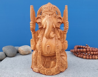 May include: A hand-carved wooden statue of the Hindu deity Ganesha. The statue is intricately detailed and features Ganesha sitting with his trunk raised and holding a sweet in his hand. The statue is painted in a warm brown color and has a traditional design.