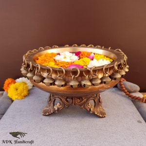 May include: A gold metal bowl with a decorative base and a ring of bells around the rim. The bowl is filled with orange and white flowers.