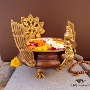 May include: A gold metal peacock-shaped bowl with a brown interior, filled with water and colourful flowers. The bowl has a peacock design on the top and a peacock tail design on the back. The bowl is sitting on a stone surface with a wooden mala necklace.