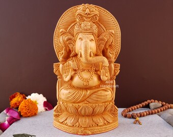 Op de afbeelding: Een handgesneden houten beeld van de hindoe-god Ganesha, afgebeeld zittend in een meditatieve houding met zijn slurf opgerold. Het beeld is ingewikkeld gedetailleerd, met een kroon, sieraden en een lotusbasis. Het beeld is geschilderd in een lichtbruine kleur.