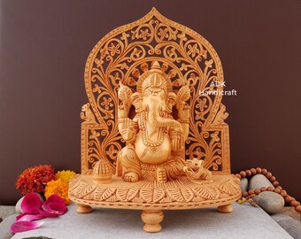 Op de afbeelding: Een handgesneden houten beeld van Ganesha, de hindoeïstische god van wijsheid en voorspoed, zittend op een troon met ingewikkelde houtsnijwerk. Het beeld is gemaakt van lichtbruin hout en heeft een gedetailleerd ontwerp. Het beeld staat op een houten voet met drie poten. Het beeld is omringd door bloemen en een houten mala.