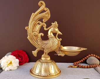 May include: A gold metal peacock-shaped oil lamp with intricate details. The lamp has a round base and a small bowl for oil. The peacock's tail is spread out behind it.