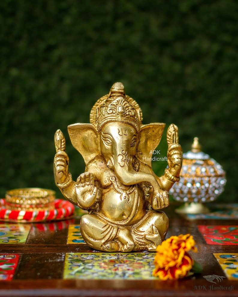 May include: A gold-colored brass statue of Ganesha, the Hindu god of beginnings, wisdom, and prosperity. The statue is sitting on a colorful wooden surface with a flower in the foreground. The statue is holding a small object in its right hand and has a trunk that is curled up.